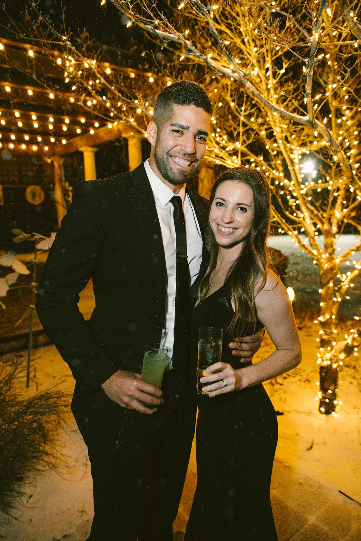 Couple in formal attire smiling under festive string lights at night.