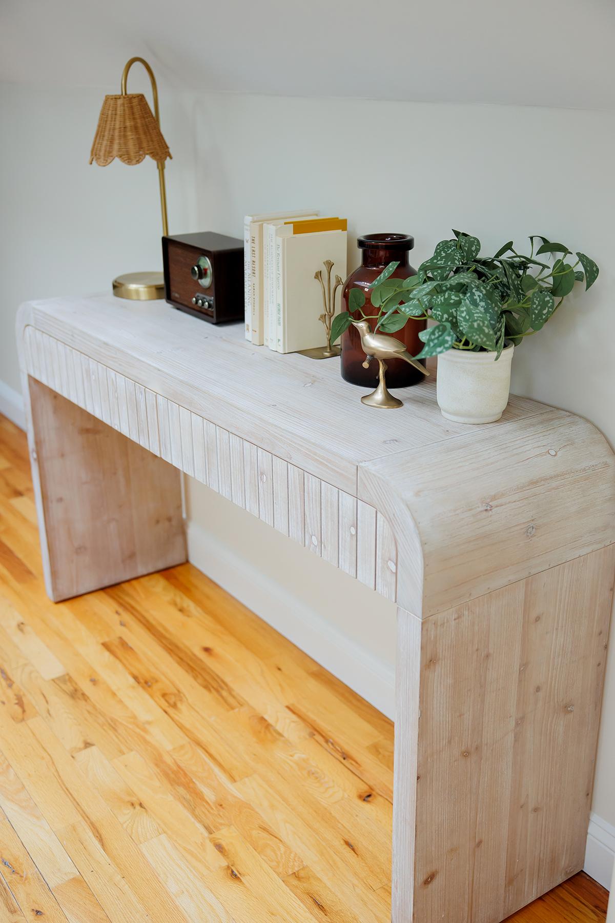 Light wooden console table with books, potted plant, lamp, and decor on a wooden floor.