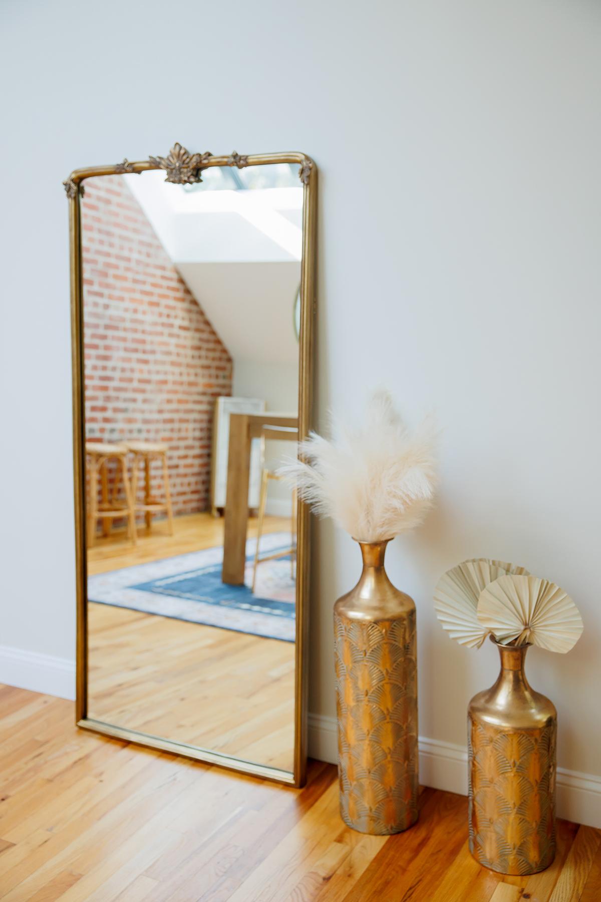 Tall mirror reflecting a brick wall and wooden stool, next to gold vases with dried plants.