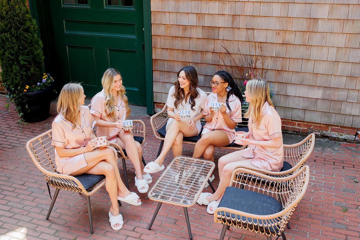 Five women in matching outfits chat and laugh on patio furniture.