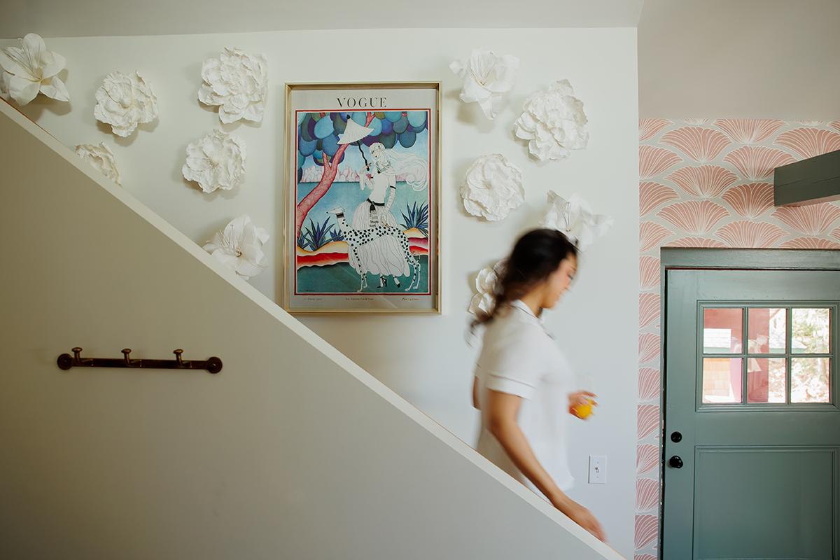 Woman descending stairs beside floral wall art and pastel door.
