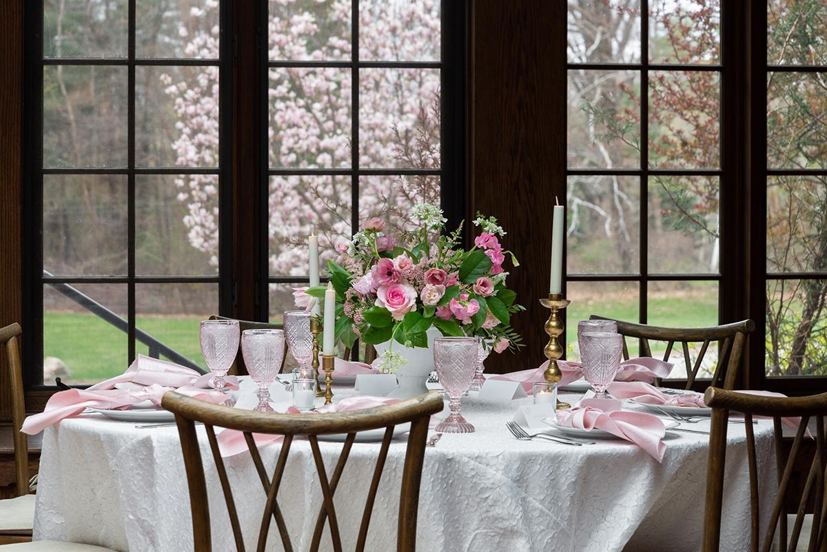 Elegant dining table with pink flowers and candles by large windows.