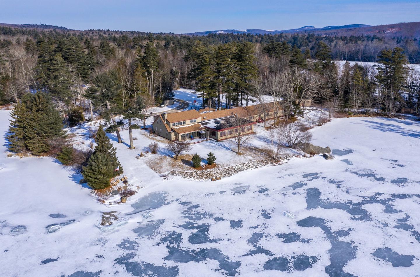 Snow-covered landscape with a house by a frozen lake, surrounded by trees.