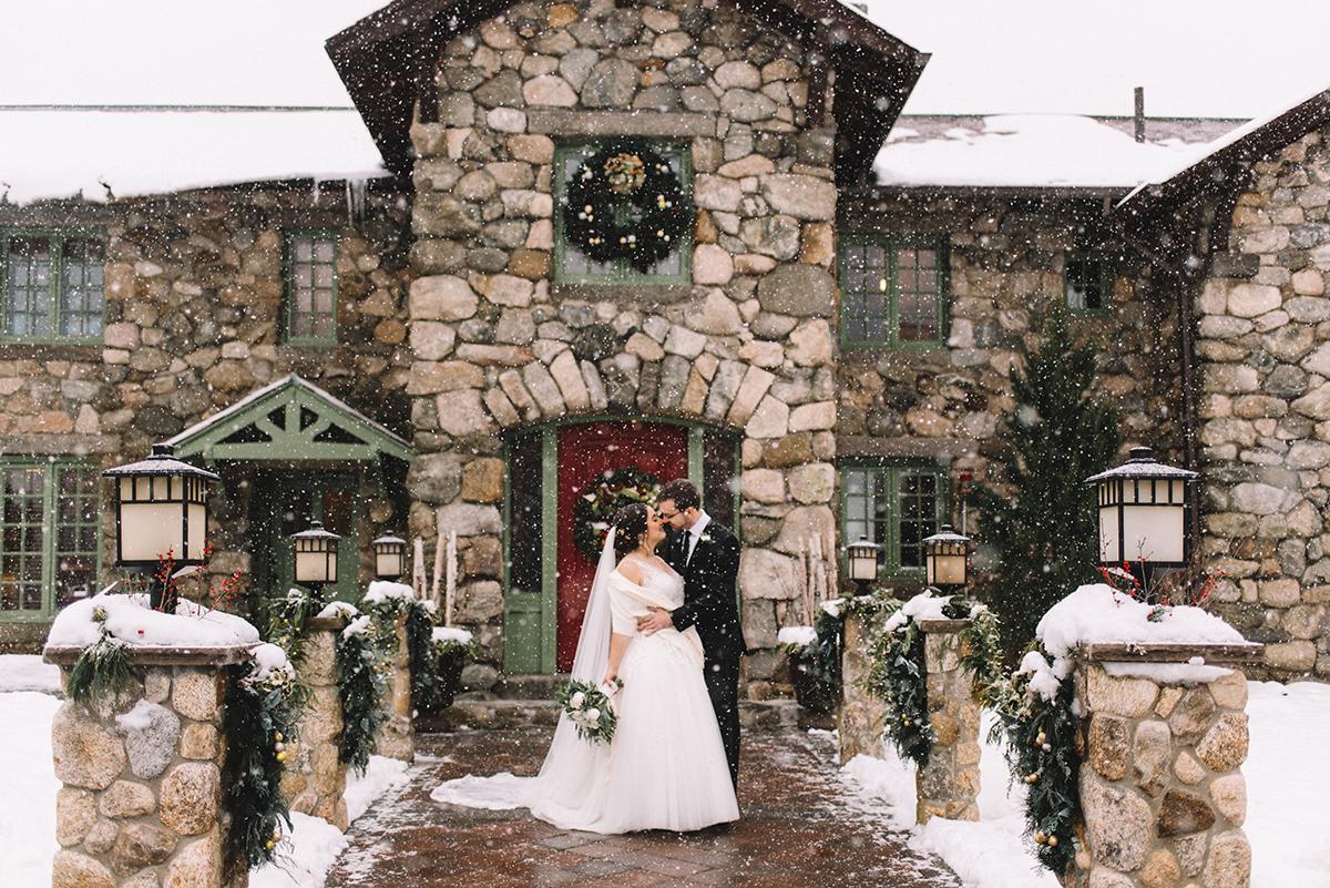 Bride and groom embrace in front of stone house with snow falling.