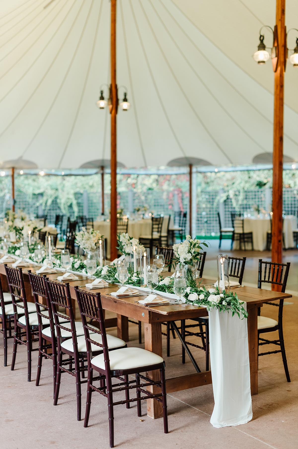 Elegant banquet table set under white tent with greenery and candles.