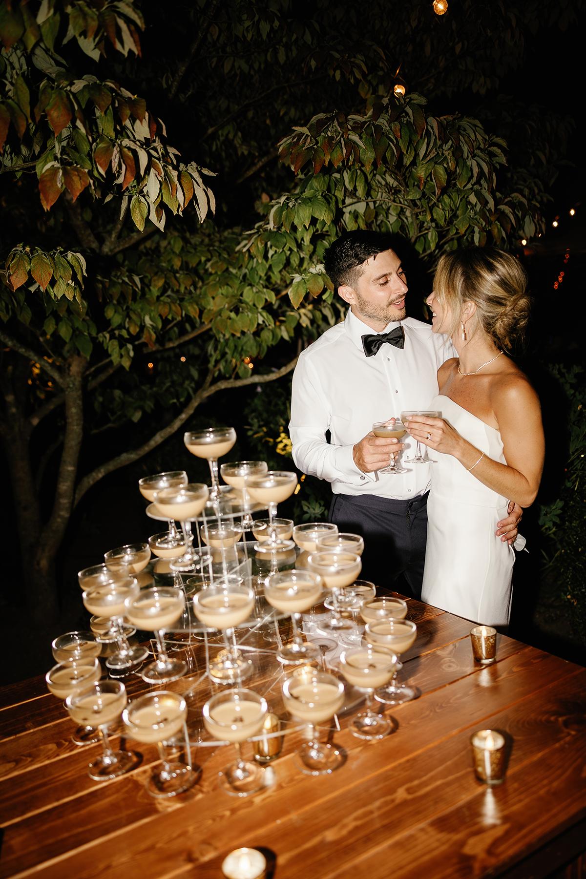Wedding couple smiling next to a pyramid of champagne glasses outdoors at night.