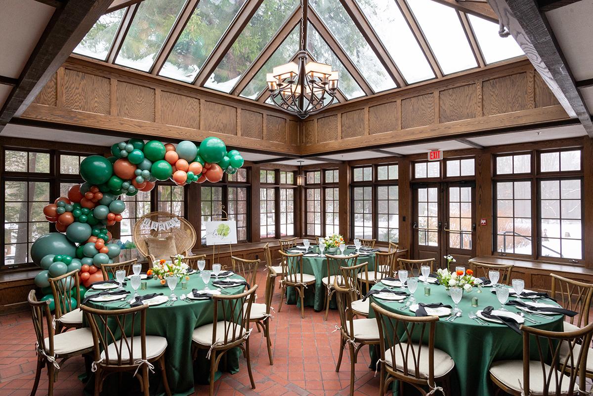 Elegant indoor event setup with green table settings and balloon decorations under a glass ceiling.