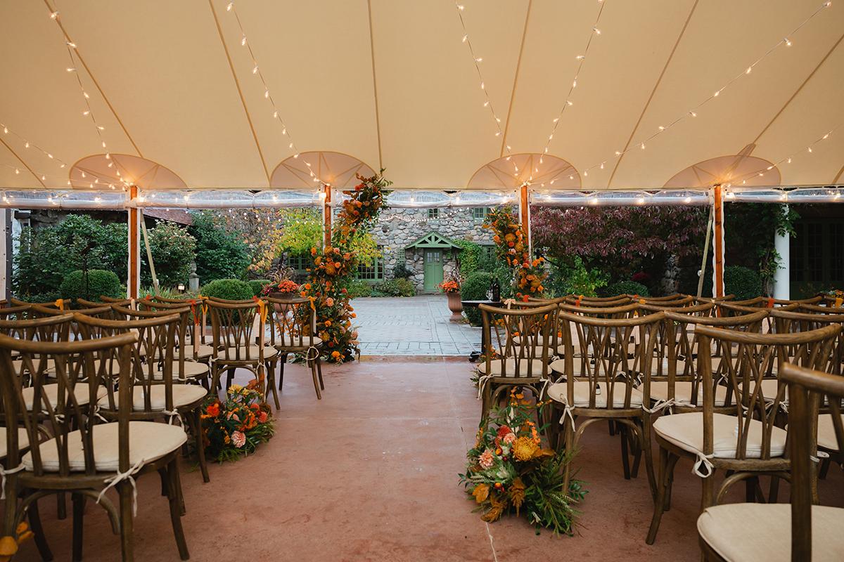 Outdoor wedding ceremony setup with floral arch and wooden chairs under a tent.