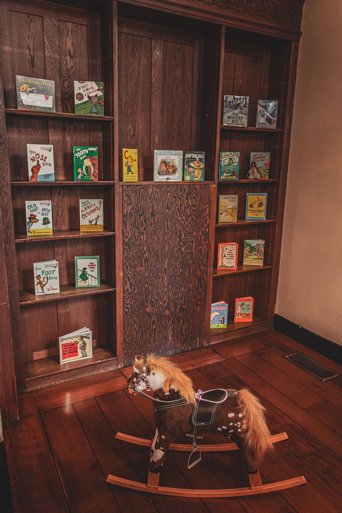 Wooden rocking horse on floor, colorful books on a wooden shelf.
