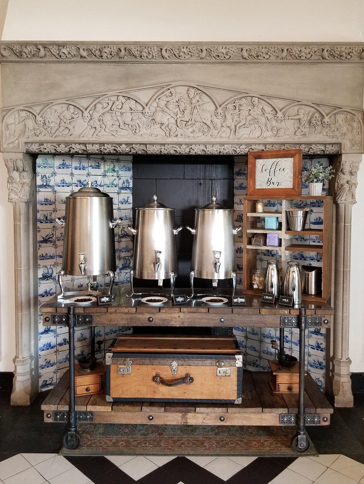 Silver coffee urns on a wooden table beneath an ornate stone mantle.