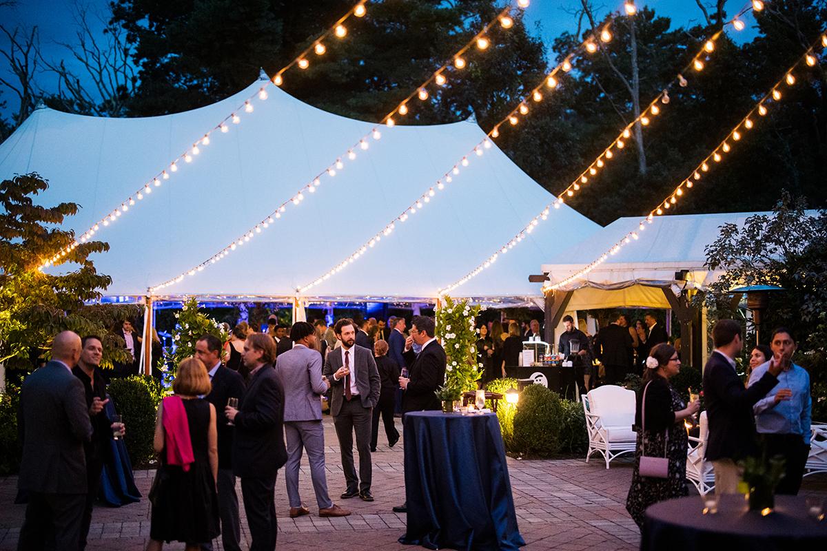 Outdoor evening event with string lights and guests mingling under a white tent.