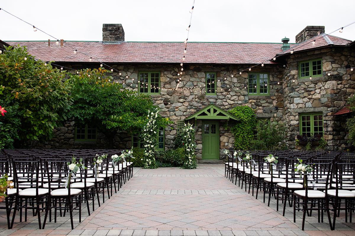 Outdoor wedding aisle facing a rustic stone building, with chairs and flowers.
