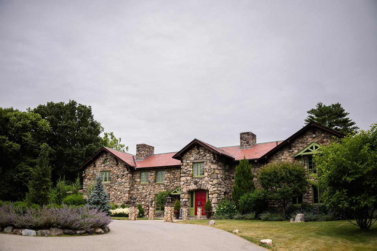 Stone house with red roof, surrounded by greenery under a cloudy sky.