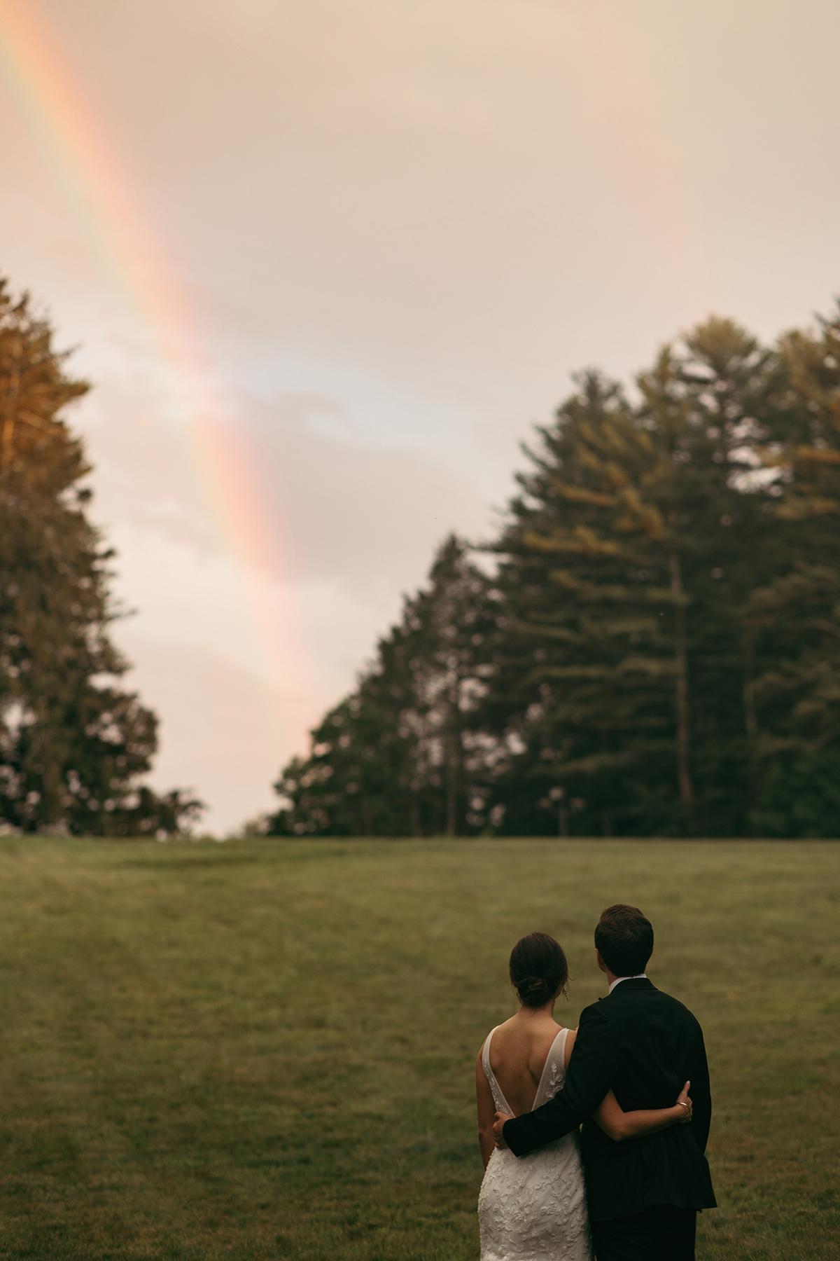 Couple embracing, facing a rainbow over a grassy field with trees.