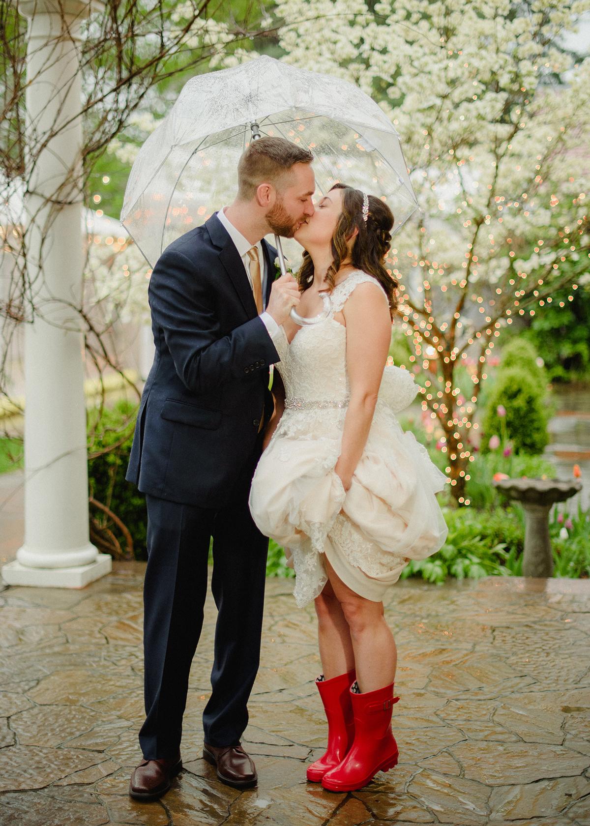 Couple kissing under umbrella, bride in red boots, garden setting.