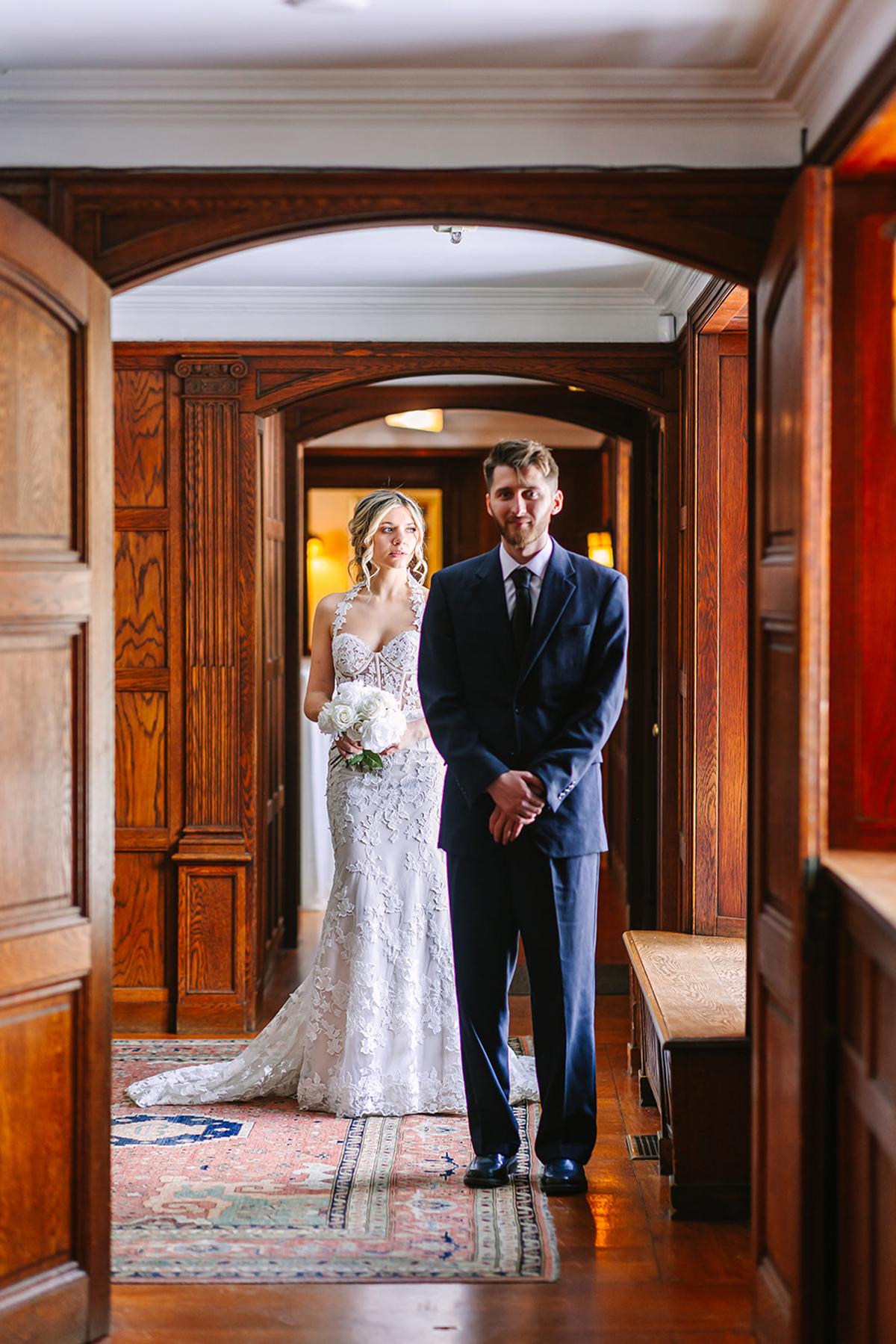Bride and man in dark suit in a wooden hallway, bride holding bouquet.
