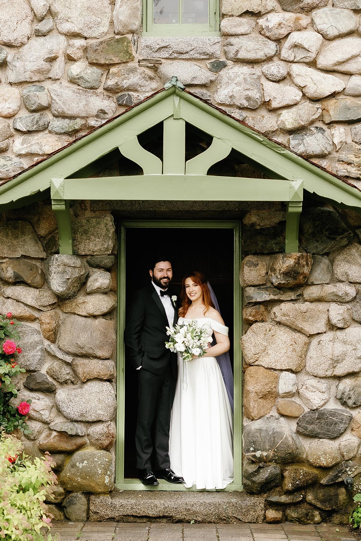 Couple in wedding attire standing in a stone doorway.