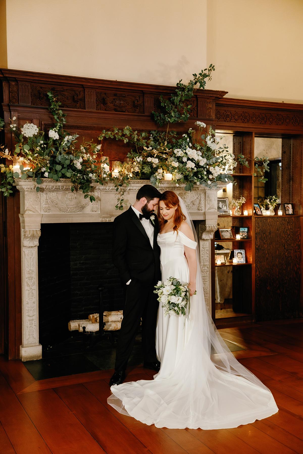 Bride and groom stand by a fireplace, surrounded by floral decorations.