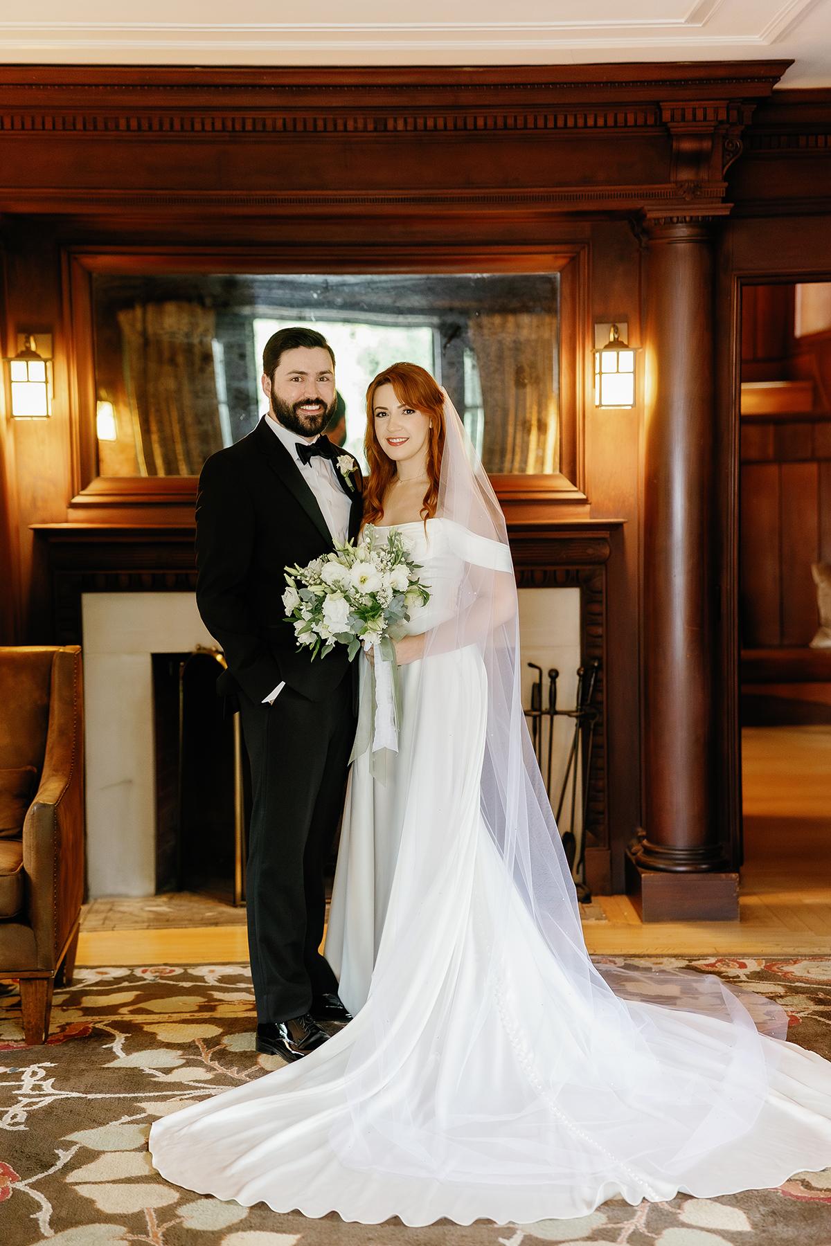 Bride and groom smiling in front of fireplace in elegant room.