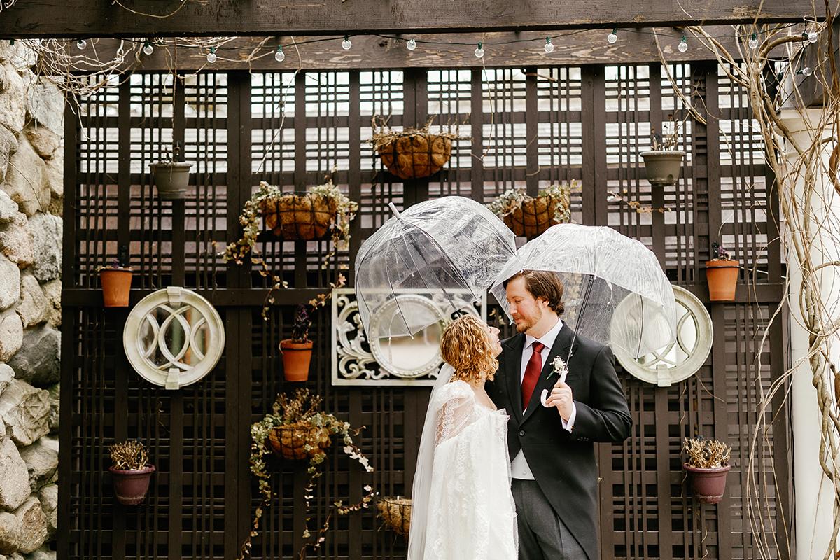Couple in wedding attire under clear umbrellas, smiling in a decorated garden setting.