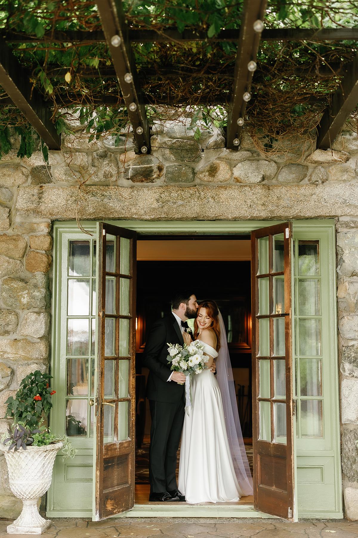 Bride and groom kissing under a stone archway with green doors.