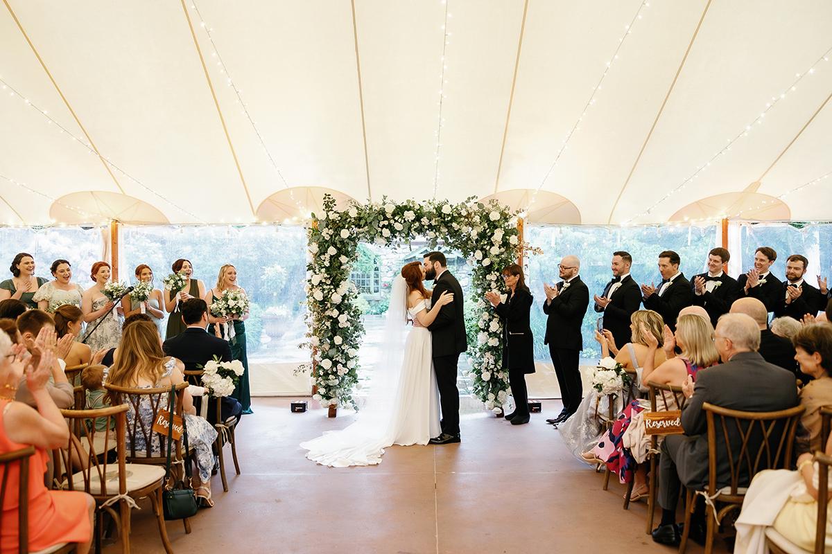 Bride and groom kiss under floral arch at a wedding ceremony.