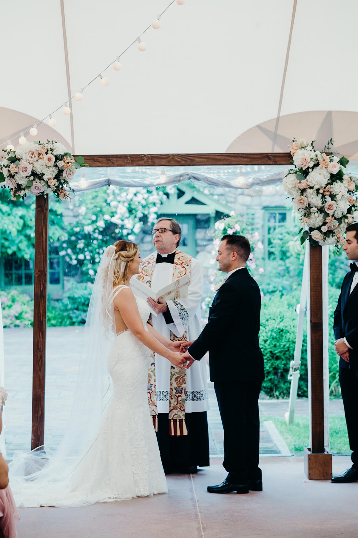 Bride and groom hold hands under floral arch during outdoor wedding ceremony.