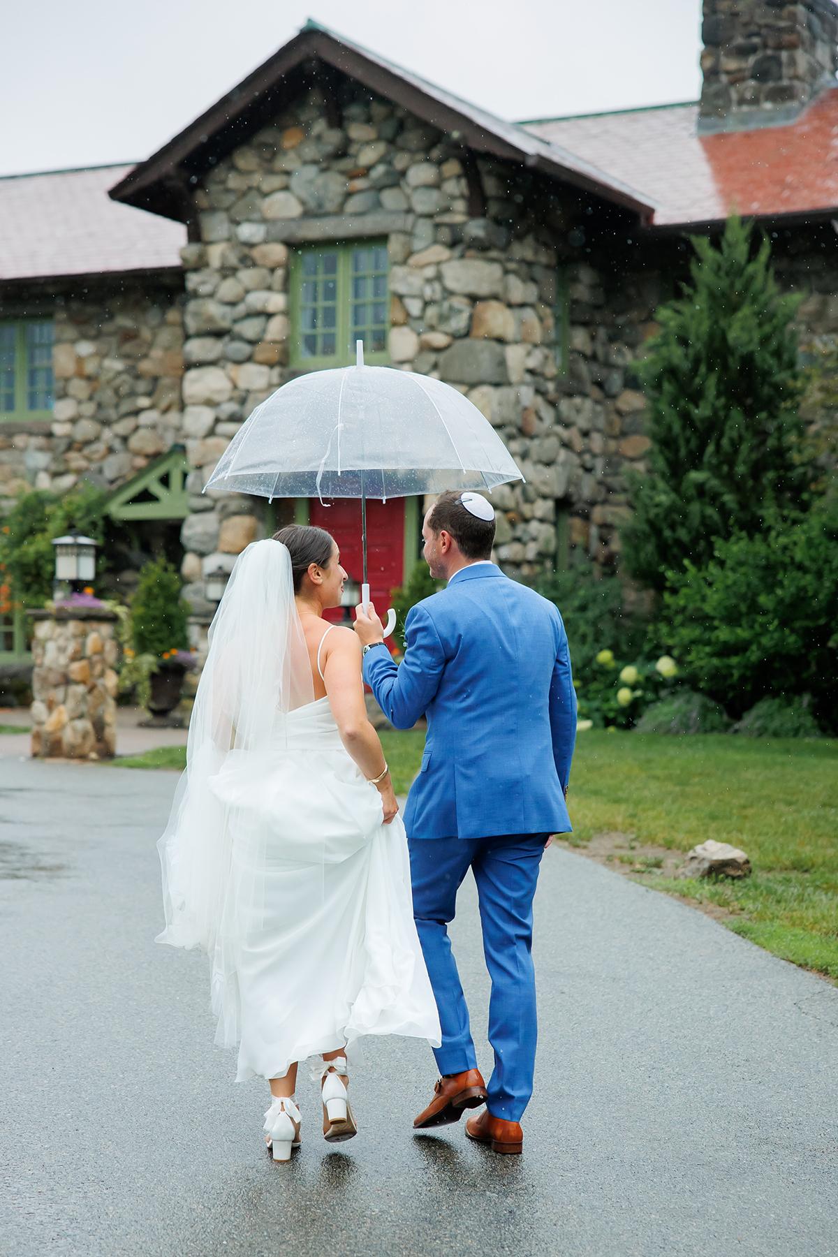 Bride and groom walk hand-in-hand under an umbrella, near a stone building.
