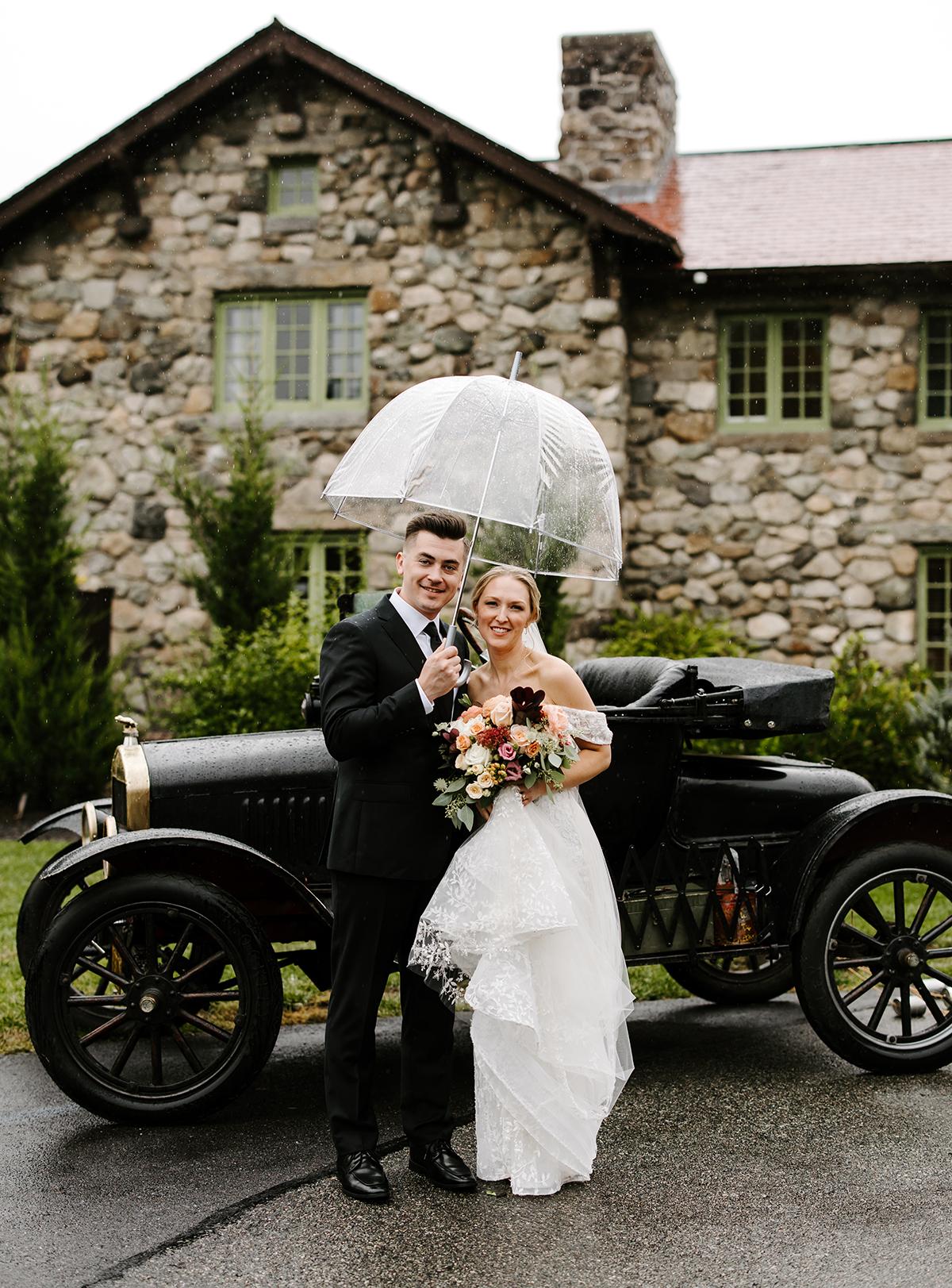 Bride and groom smile under an umbrella by a vintage car and stone building.