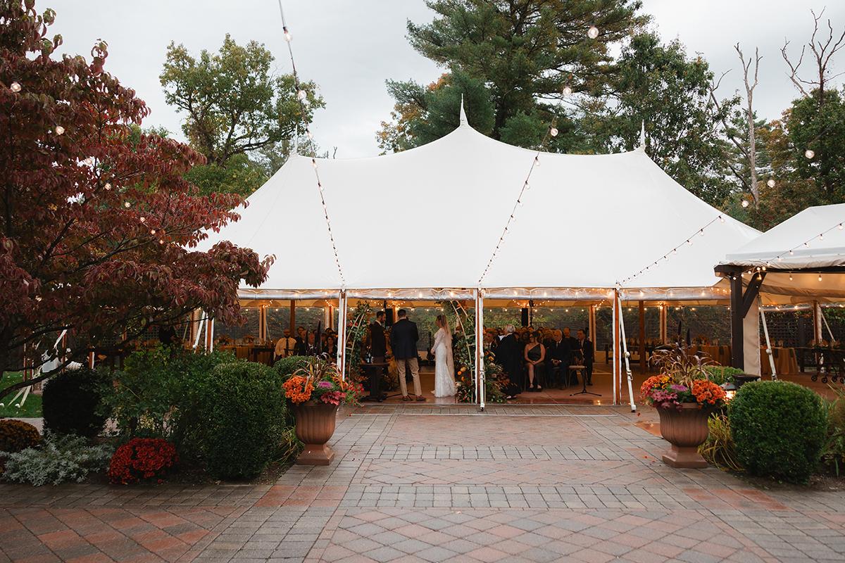 Wedding tent with guests, surrounded by fall foliage and potted plants.