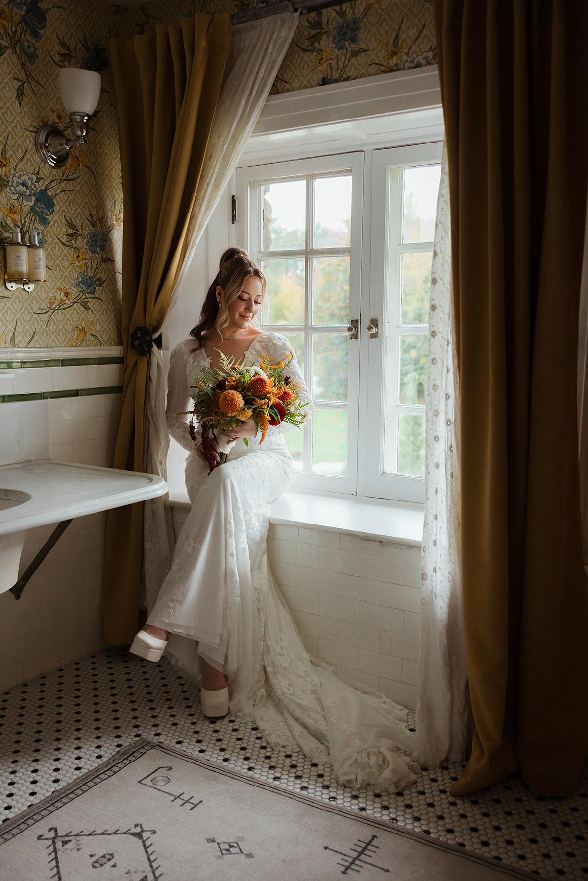 Bride in a white dress holding flowers, sitting by a window in a floral-themed room.