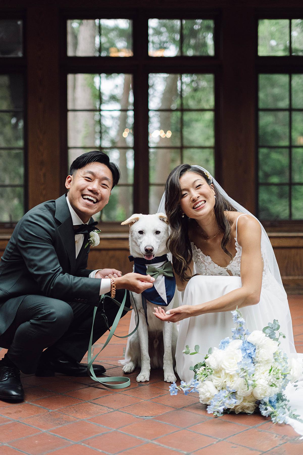 Bride and groom smiling with their dog, indoors, bouquet nearby.