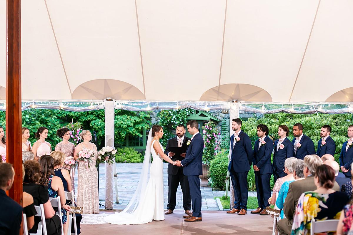 Bride and groom holding hands under a tent, surrounded by their wedding party.