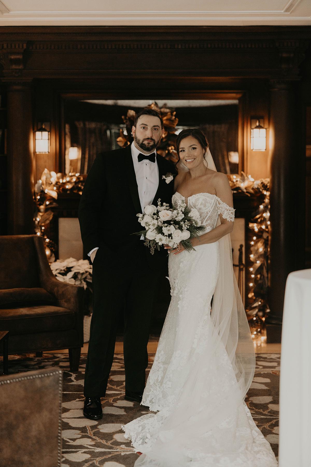 Couple in wedding attire standing indoors, warmly lit decor in background.