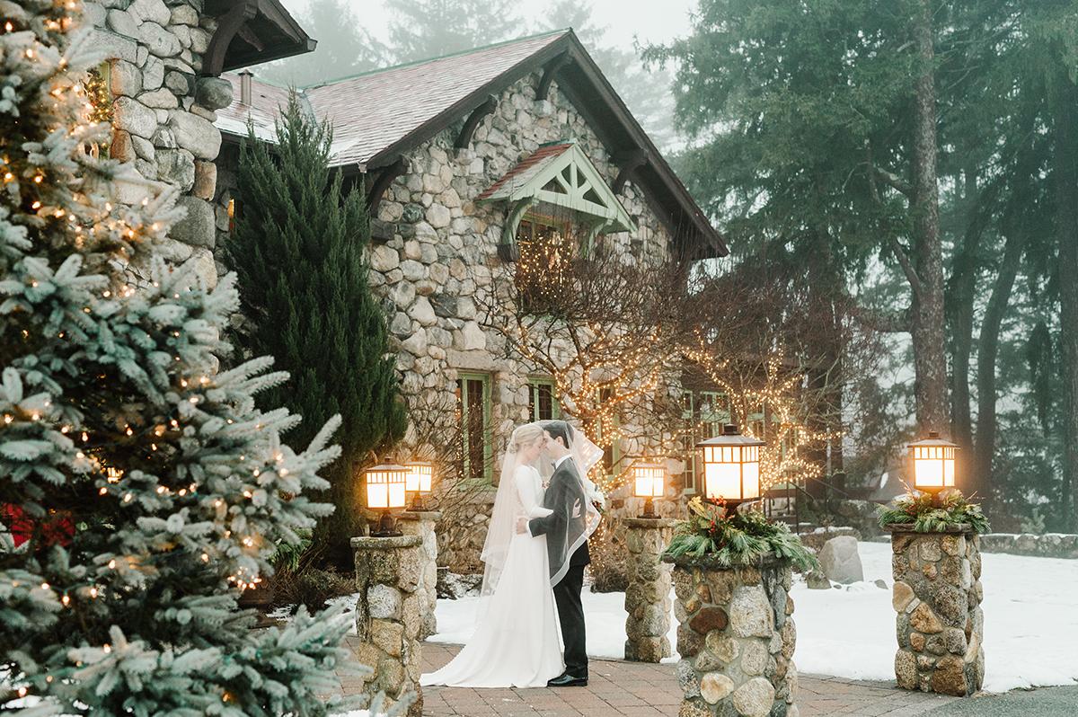 Bride and groom in winter setting by stone house, surrounded by snowy trees.