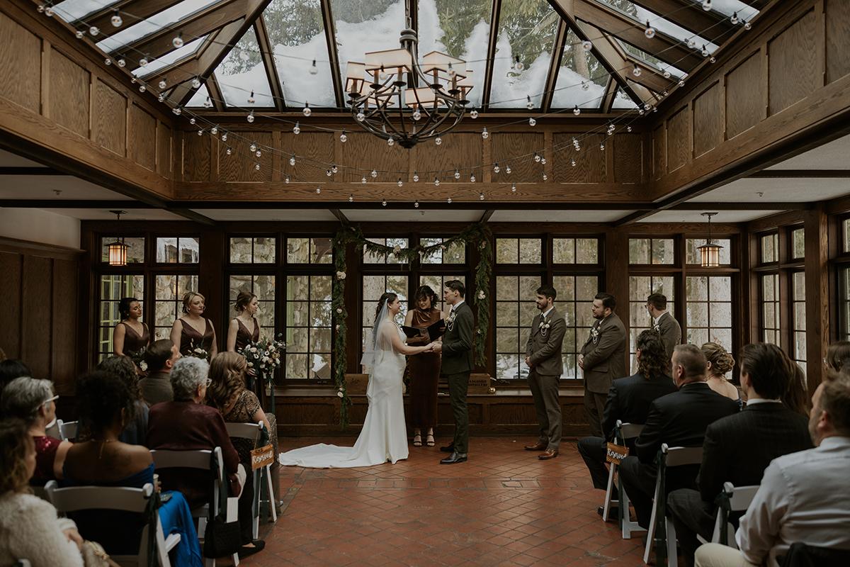 Indoor wedding ceremony with bride and groom under a decorated arch. Guests seated in rows.