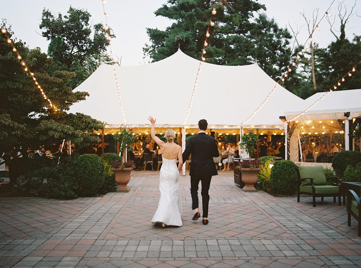 Bride and groom walking towards tent with string lights in evening garden setting.