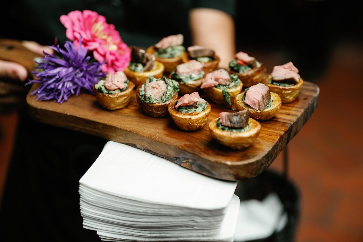 Mini tarts topped with meat on a wooden tray decorated with pink and purple flowers.