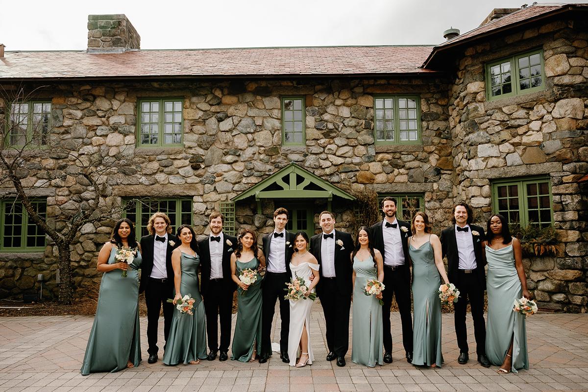 Bridal party in formal attire poses in front of a stone building.