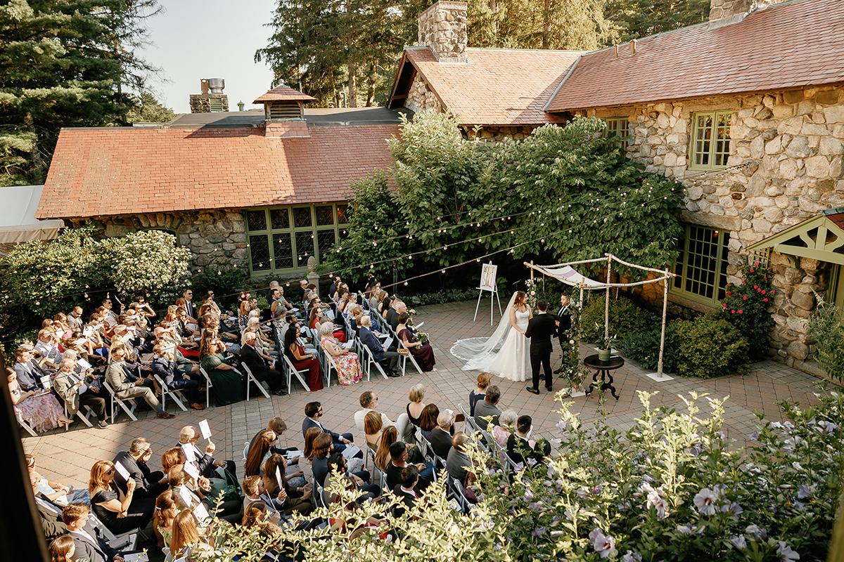 Wedding ceremony outdoors by stone cottages, guests seated, greenery surrounds.