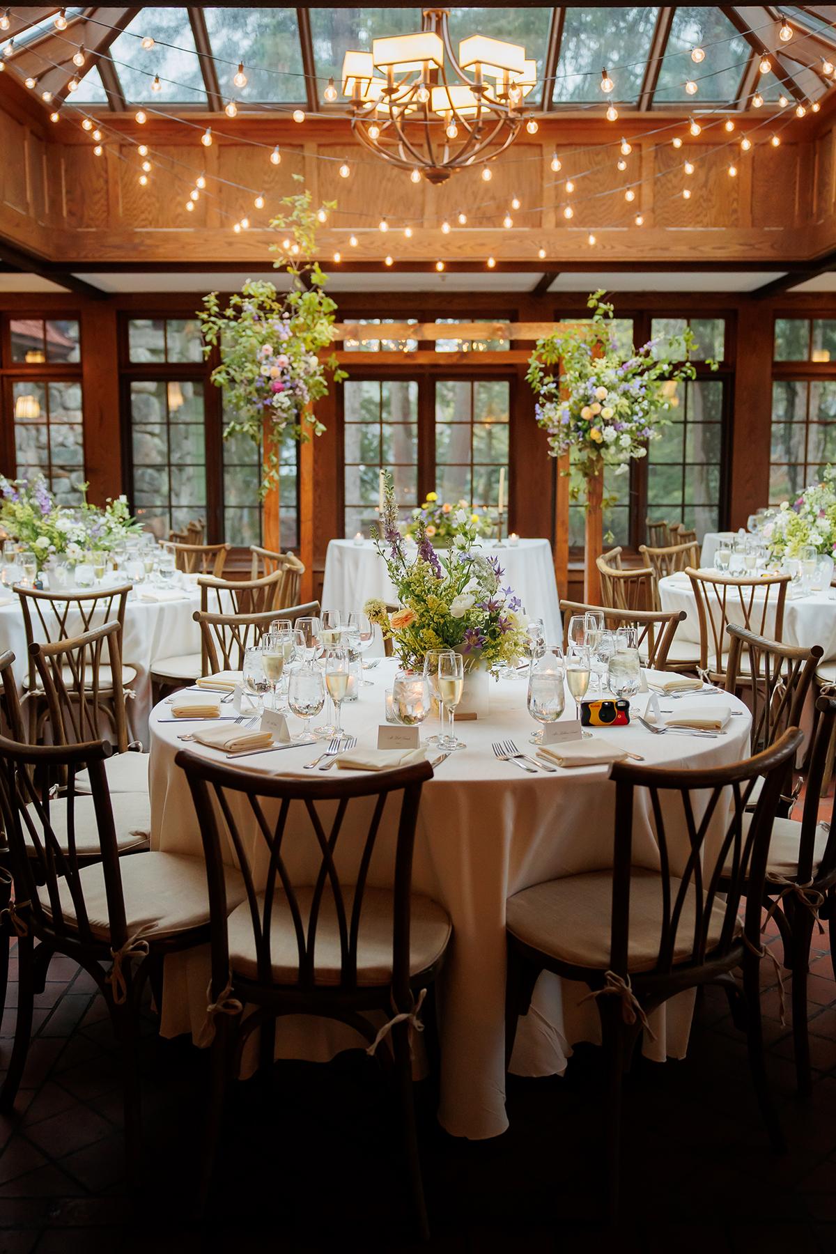 Elegant dining room with round tables, flowers, twinkling lights, and large windows.