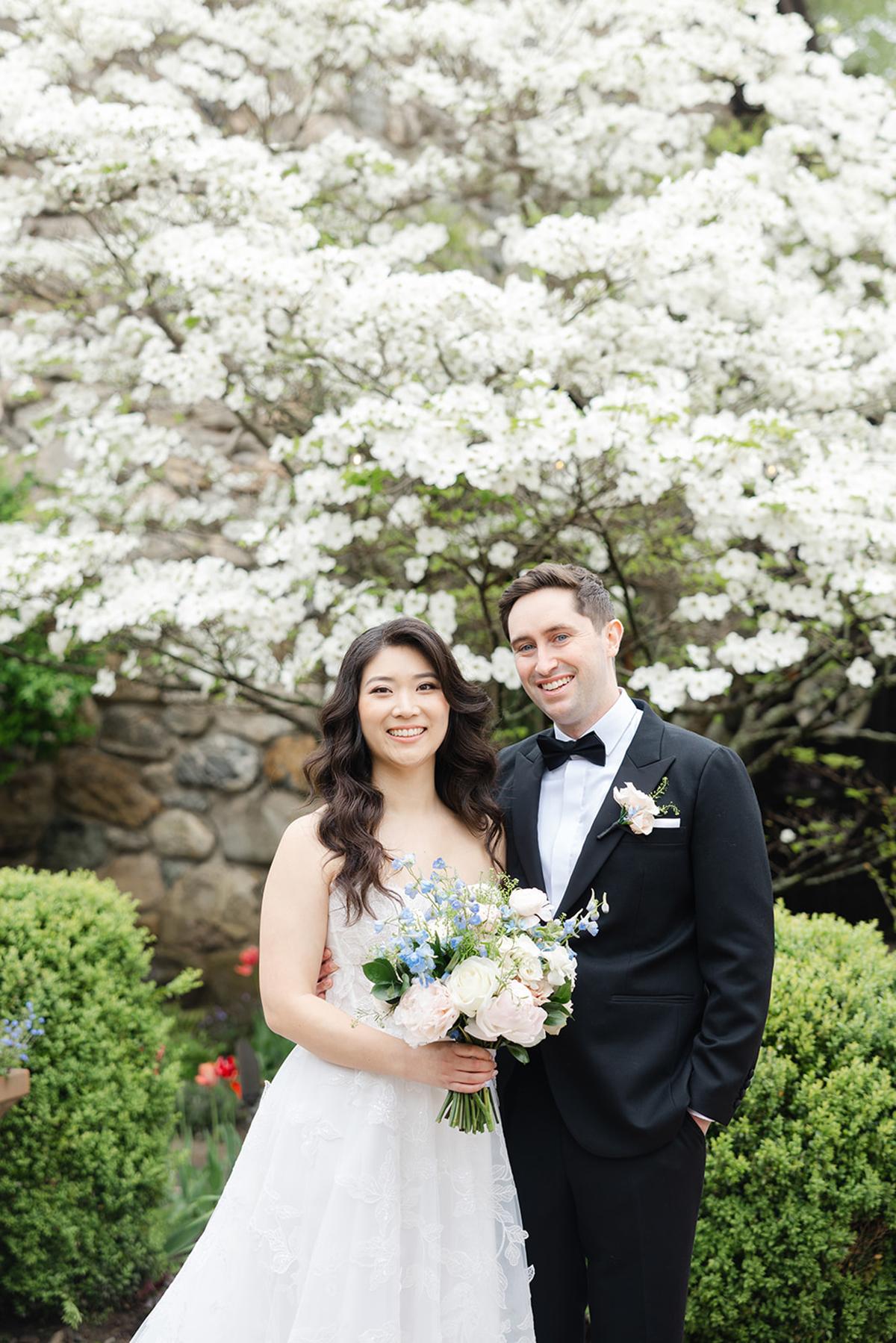 Bride and groom smiling under blooming white tree, holding a bouquet.