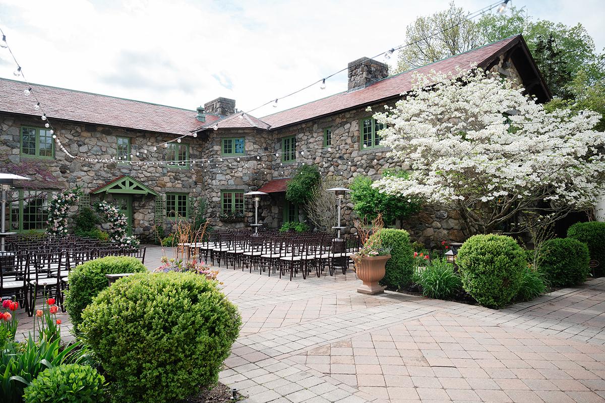 Stone building with red roof, surrounded by blooming trees and bushes on a paved patio.
