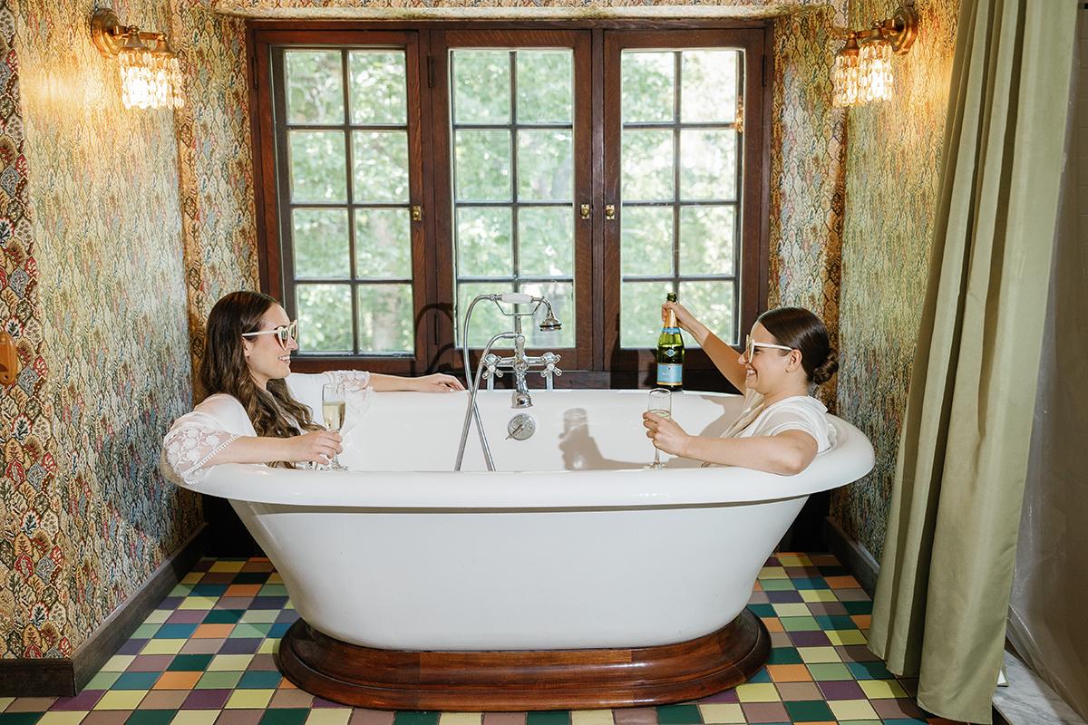 Two people relaxing in a bathtub with drinks, surrounded by patterned walls.