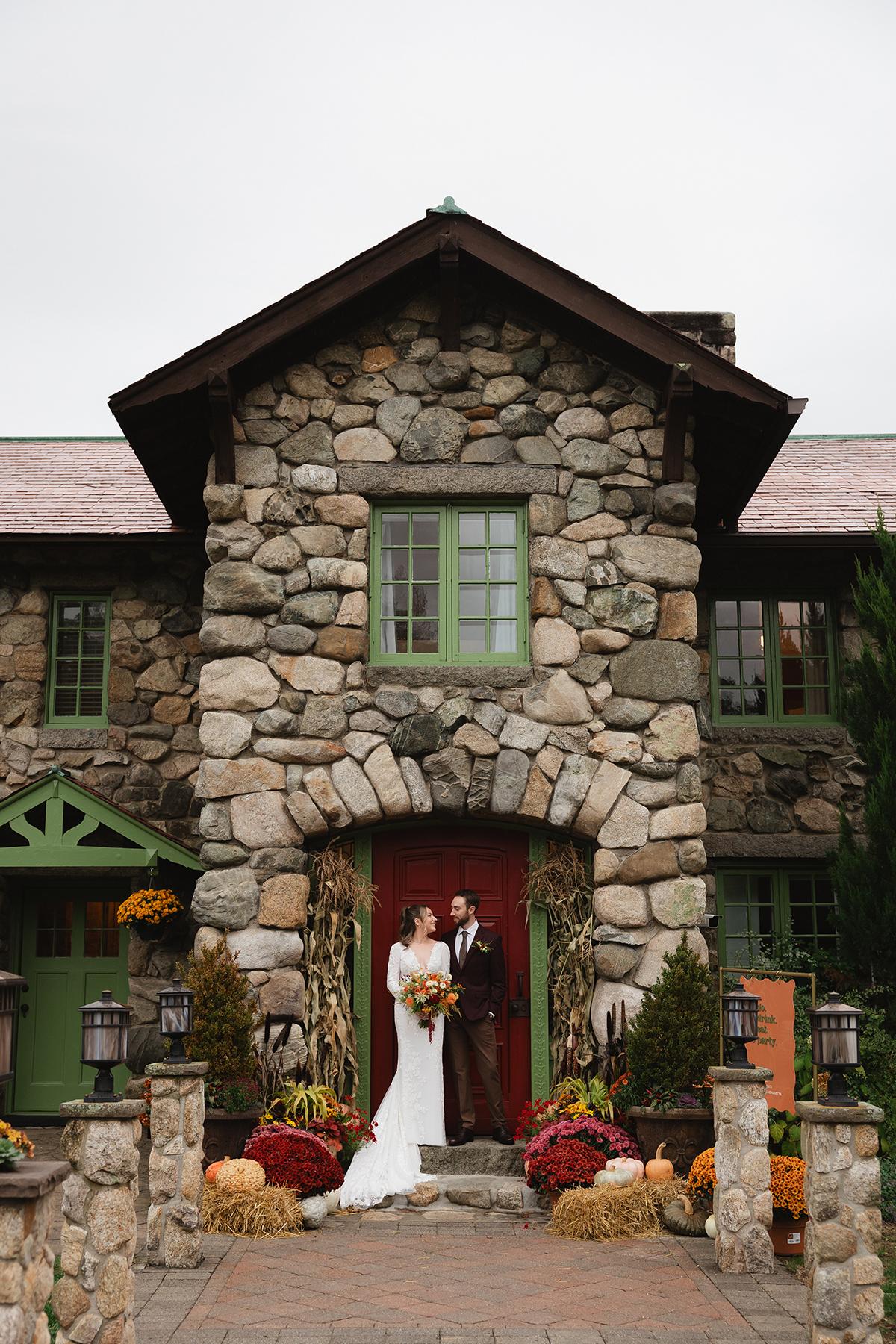 Bride and groom stand in front of a stone house, surrounded by autumn decorations.