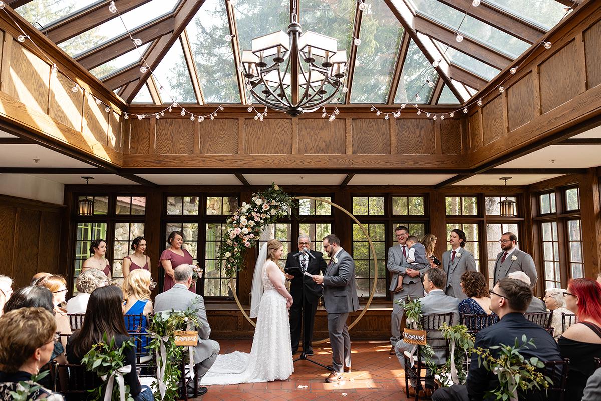 Wedding ceremony in a sunlit hall with a glass ceiling and guests seated.