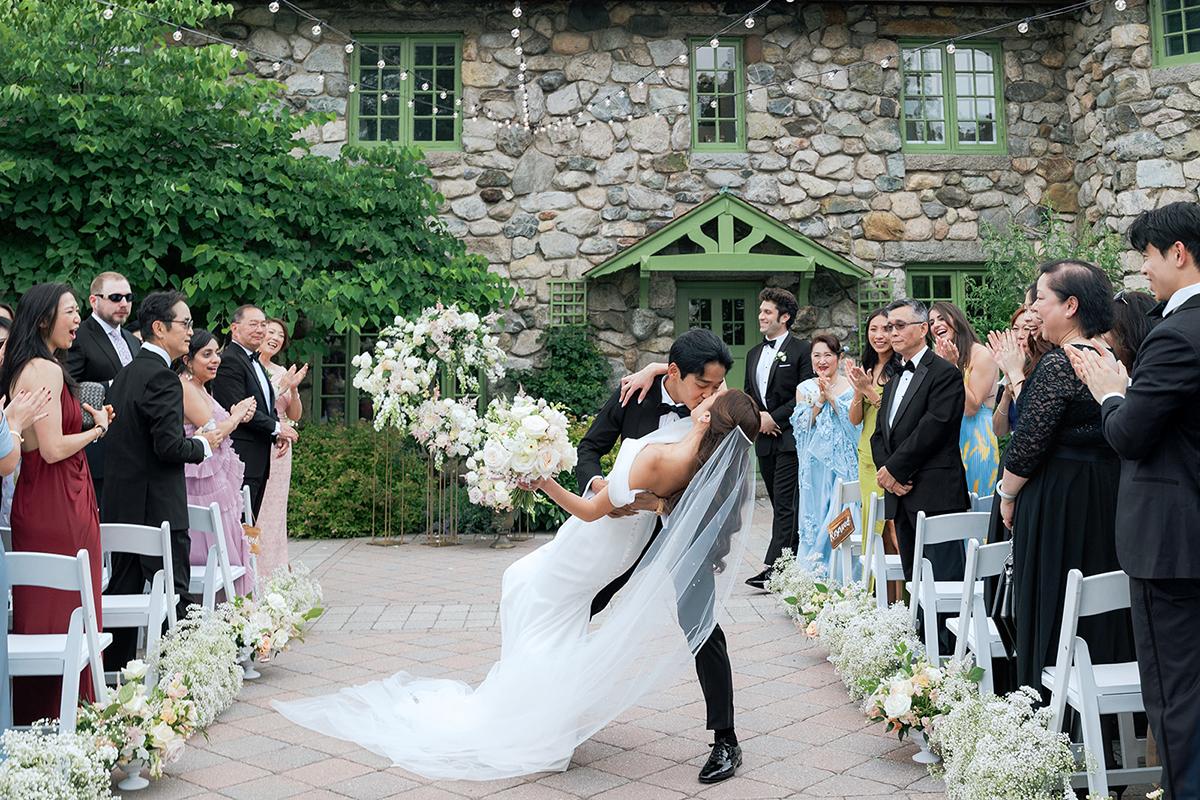 Bride and groom kissing amid clapping guests at outdoor wedding ceremony.