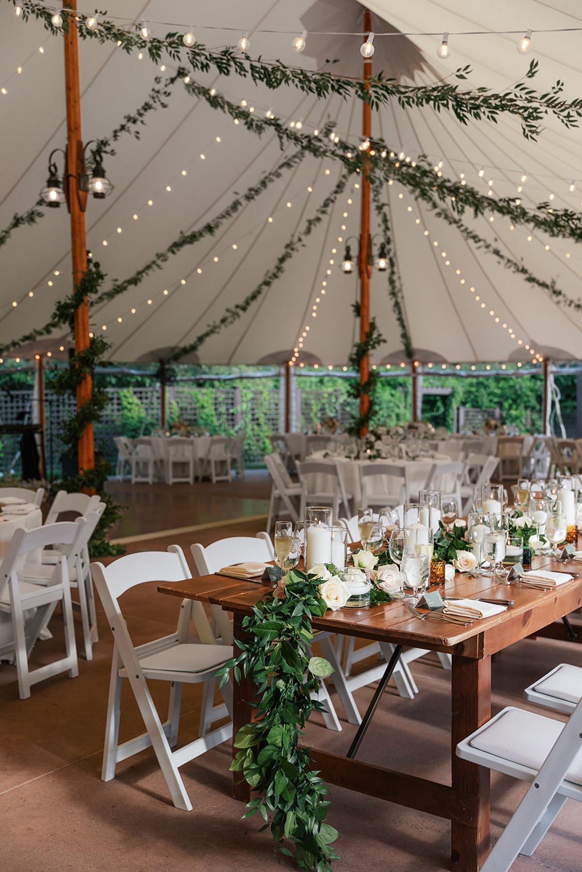 Wedding tent with white chairs, wooden table, and greenery decorations.