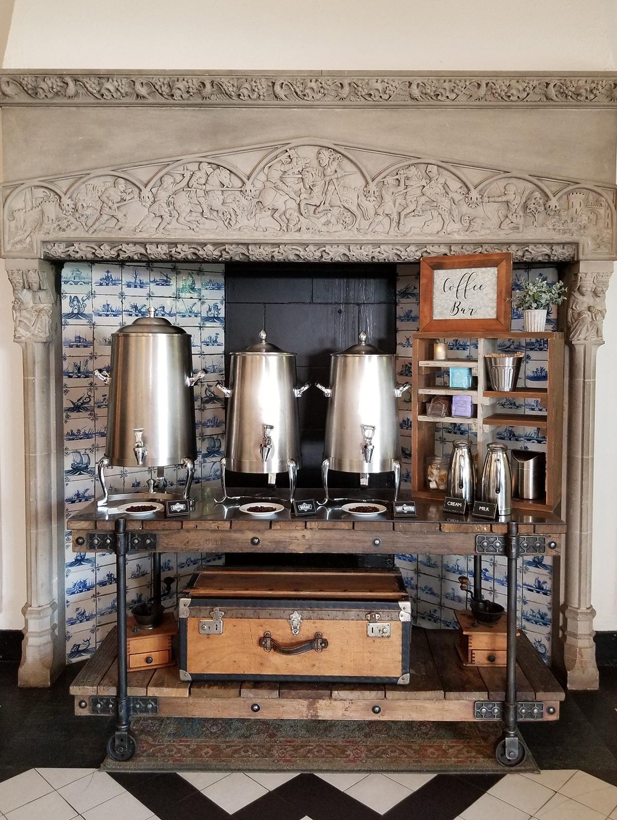 Three silver coffee urns on a decorative mantel with a vintage suitcase below.
