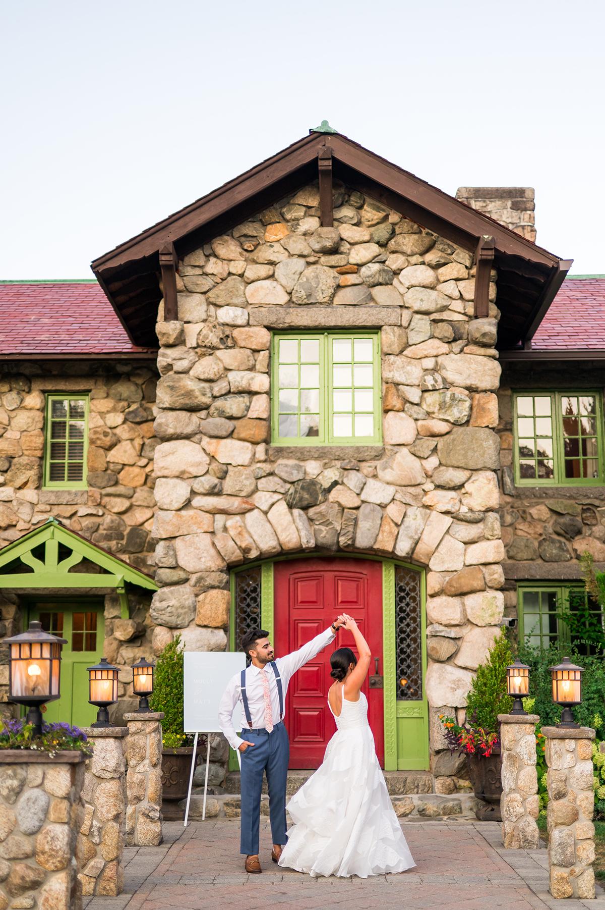 Bride and groom dancing in front of a stone house with red and green accents.